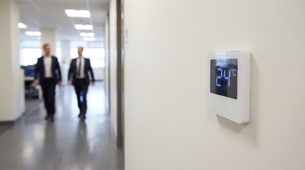 Digital wall thermostat set to 24 degrees Celsius in an office corridor with two staff members walking past in the blurred background