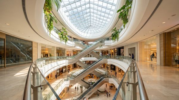 Bright multi-storey shopping mall atrium in Malaysia with a glass skylight, criss-crossing escalators and shoppers on the concourse