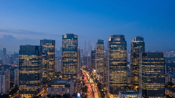 Kuala Lumpur office towers at dusk with floors of interior lights switched on, evening traffic light trails on the highway below, illustrating peak-hour electricity demand
